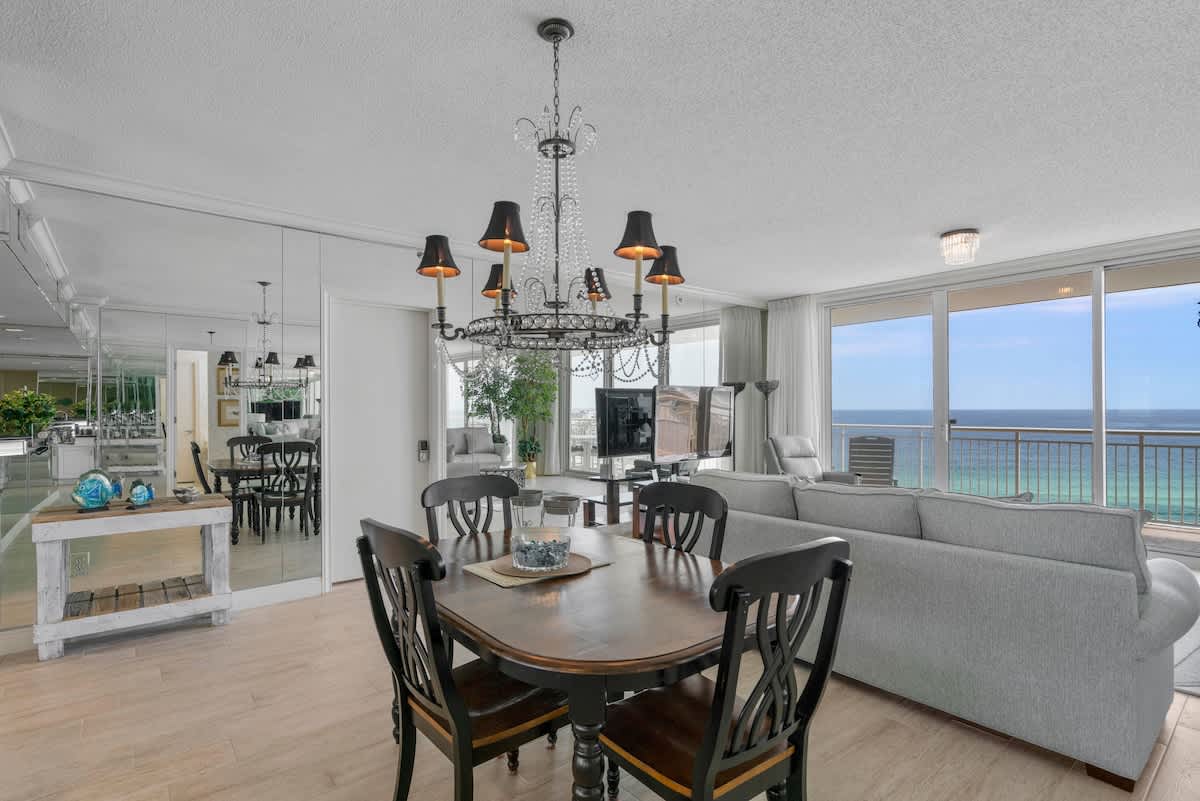 A classic dining area with a dark wood oval table and black chairs, centered under an ornate black and crystal chandelier. Mirrored walls reflect the open-concept space, including a glimpse of the ocean view.