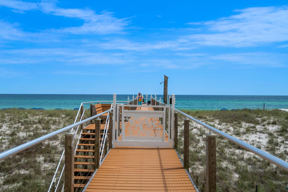 A wooden boardwalk with metal railings extends over sandy dunes towards the vast, calm blue ocean under a bright sky. A white gate with a decorative pattern marks the entrance to the beach.