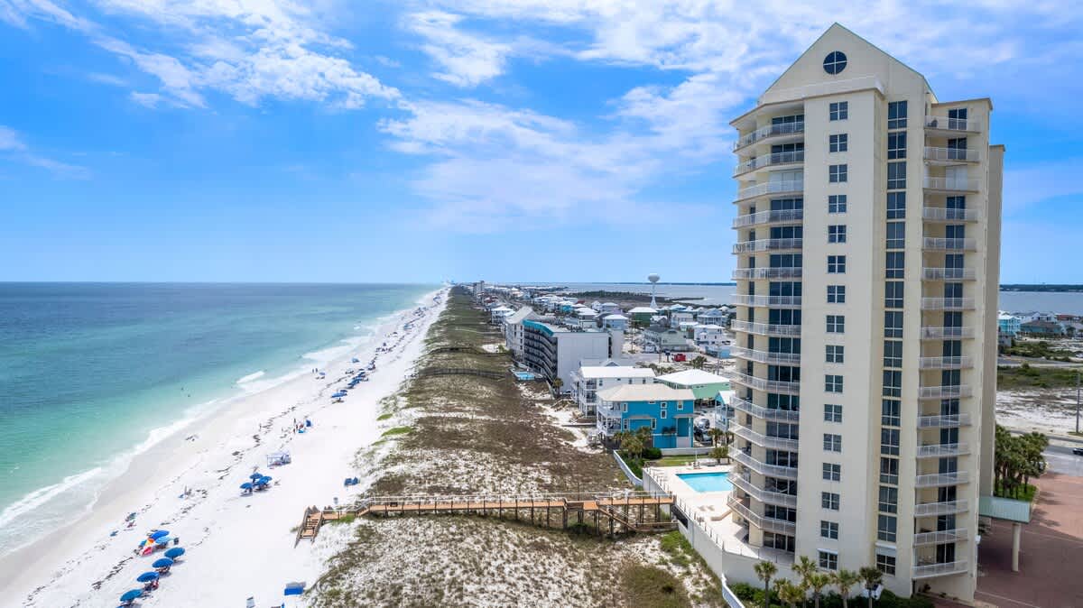 Aerial view of a tall, light-colored condominium building with a green roof, situated directly on a long stretch of white sand beach with turquoise ocean water. A wooden boardwalk leads to the beach, and other coastal buildings are visible in the distance.