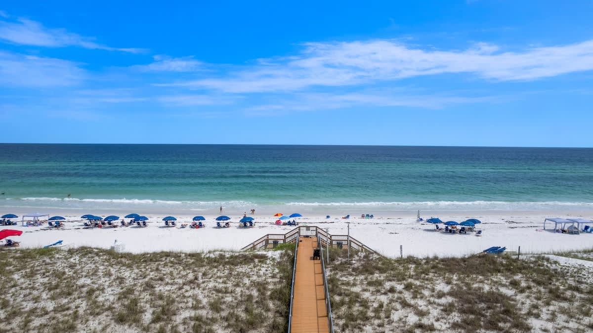 Wide aerial view of a pristine white sand beach dotted with blue beach umbrellas and chairs, leading to clear turquoise ocean waters with gentle waves. A wooden boardwalk provides access from the sandy dunes.