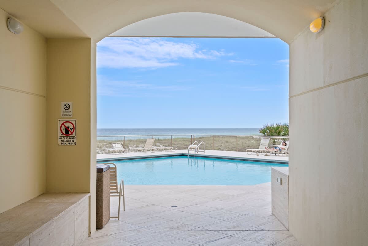 A covered archway entrance leading to an outdoor pool area, with the sparkling blue pool and the vast ocean visible in the distance under a bright sky.