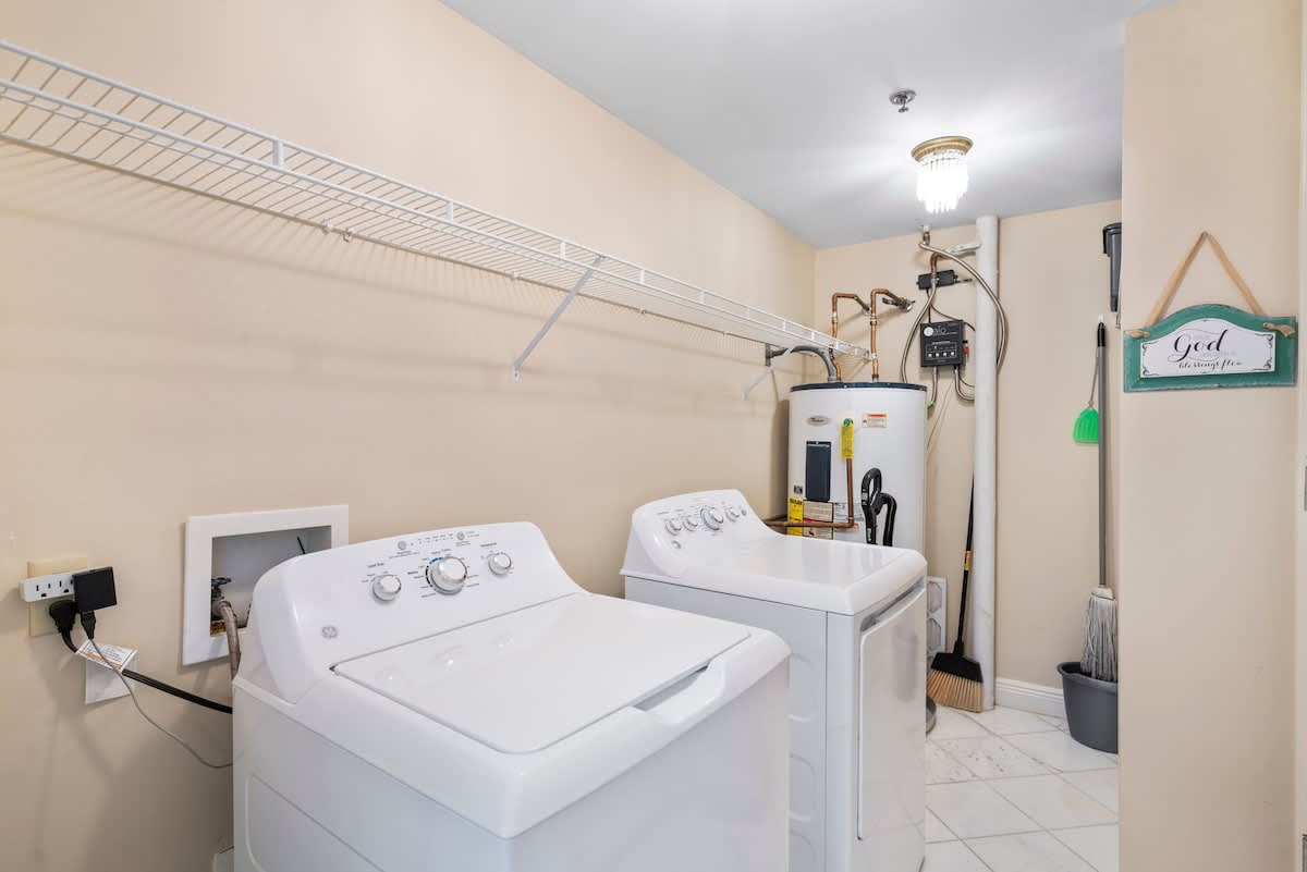 A functional laundry room equipped with a white top-loading washing machine and dryer, a utility sink, and overhead wire shelving. The walls are a light beige color.