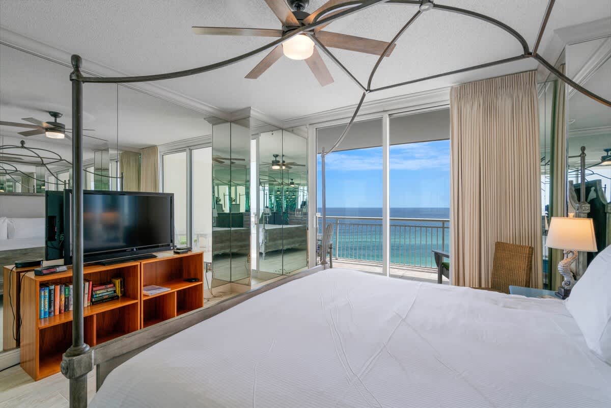 This bedroom showcases a metal canopy bed with white linens, mirrored walls, and a ceiling fan, with a large sliding glass door leading to a balcony with panoramic ocean views. A TV is placed on a wooden stand.