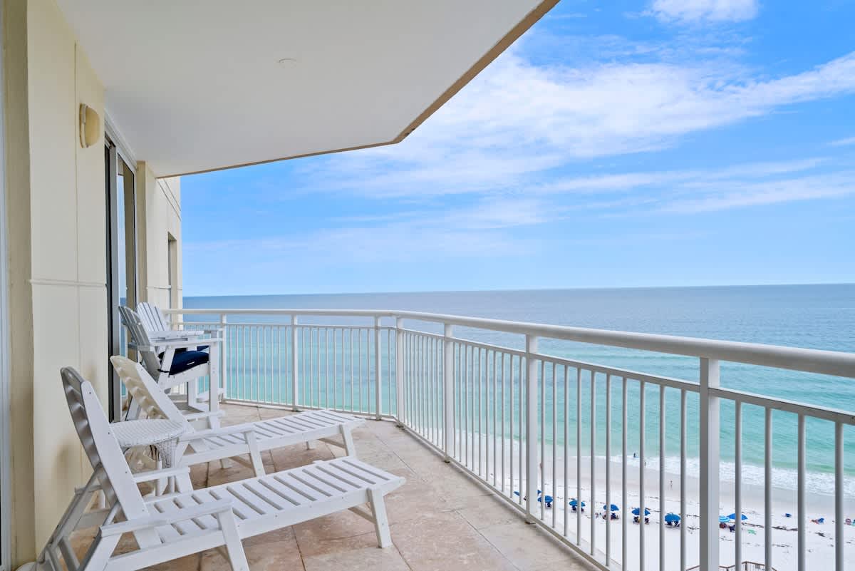 A wide balcony overlooking the ocean and beach, furnished with white lounge chairs and offering unobstructed views of the water and horizon under a bright blue sky.