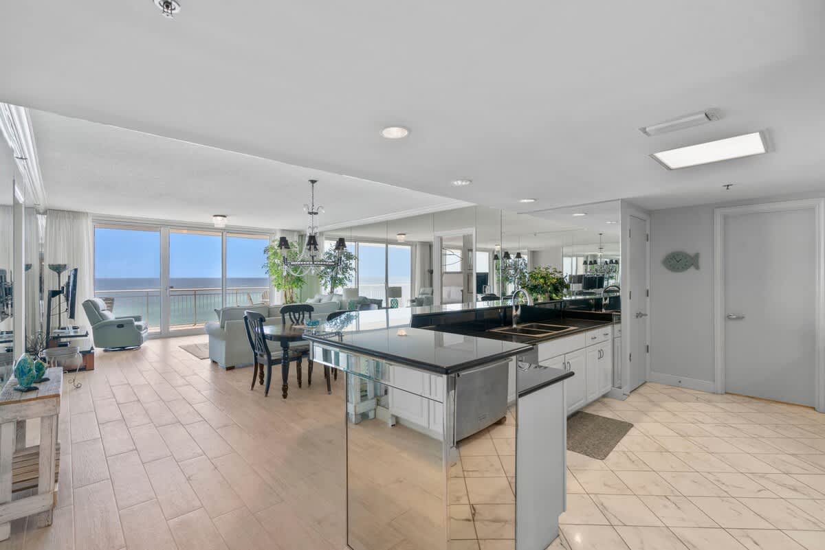 A wide view of the open-concept living space, showing the kitchen with its mirrored island and black countertops, flowing into the dining and living areas with ocean views. Light tile flooring extends throughout.