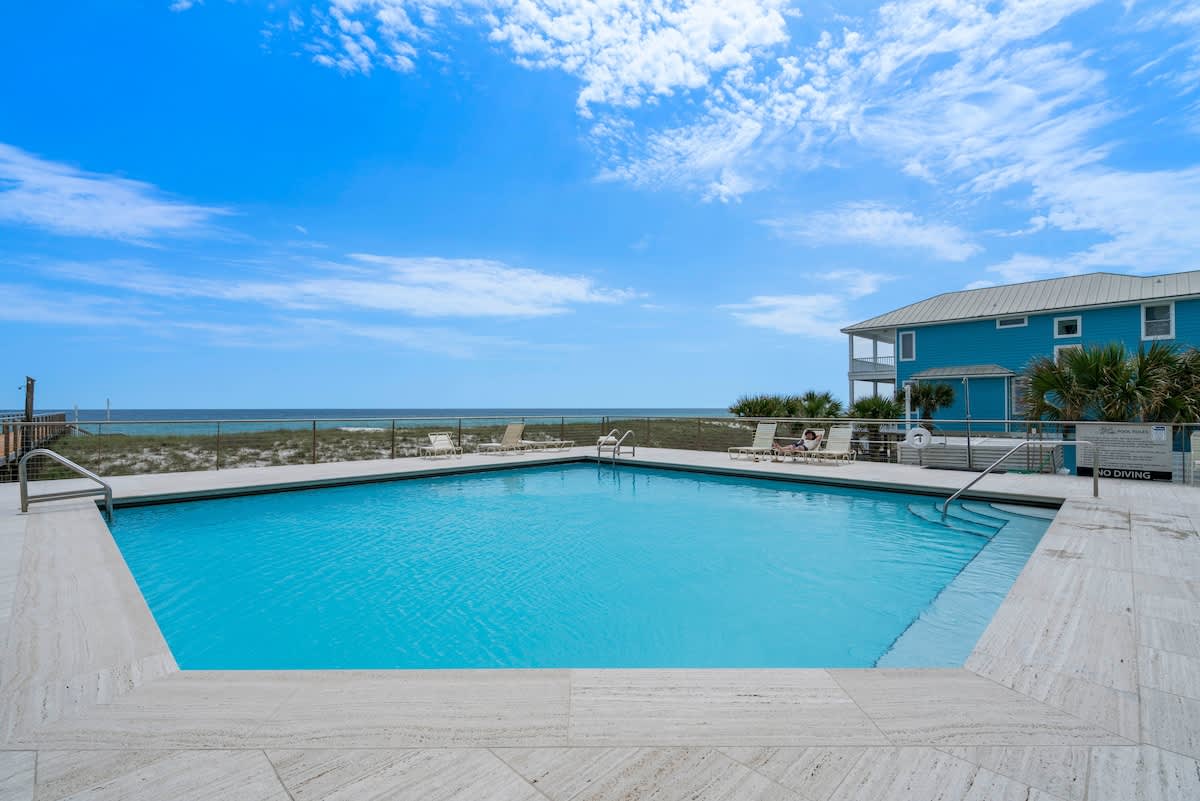 A spacious outdoor swimming pool area with a light-colored deck, offering expansive views of the sandy beach, the ocean, and a nearby blue-sided building under a clear blue sky.