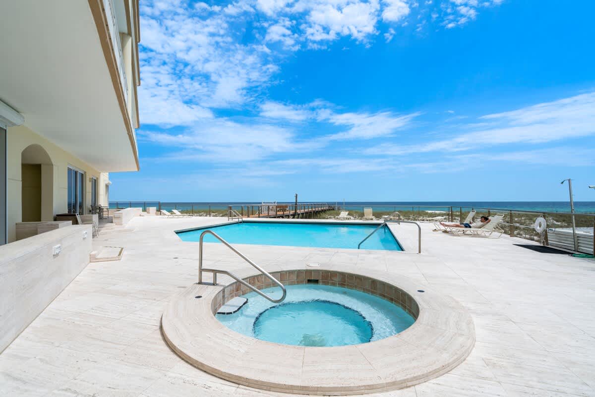 An inviting outdoor pool area featuring a rectangular swimming pool and a circular hot tub, both surrounded by a light-colored deck, with the ocean visible in the background.
