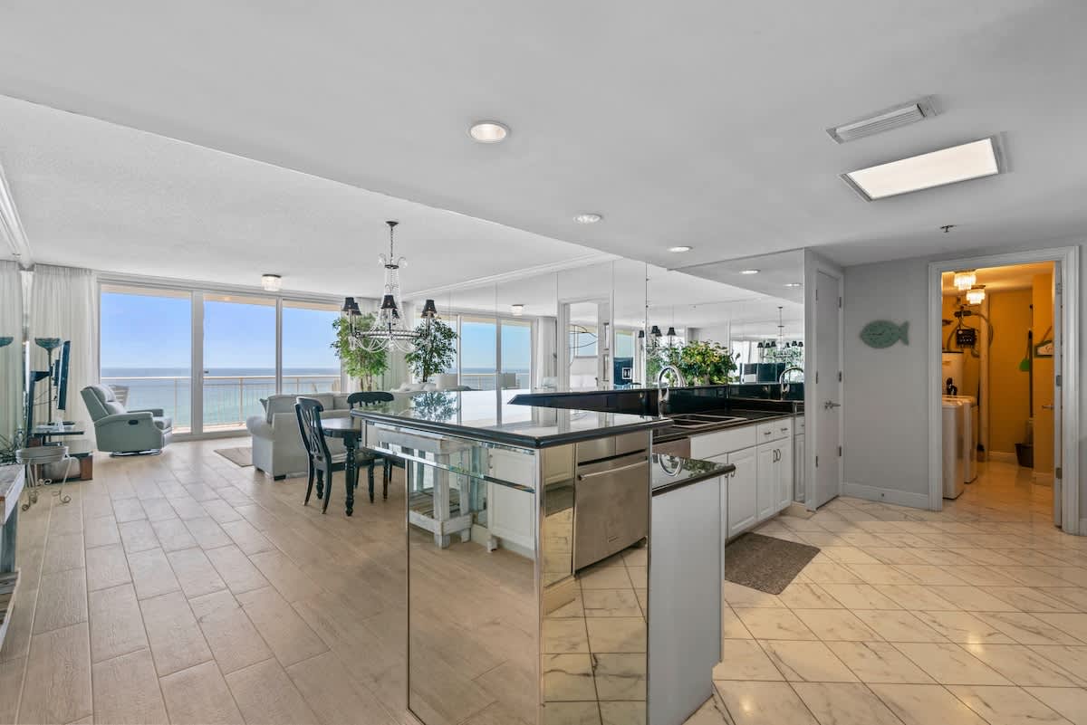 An expansive view of the open-plan living area, showcasing the kitchen with its mirrored island and black countertops, the dining space, and the living room with ocean views through large glass doors.