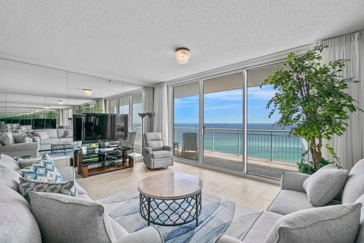 A spacious living room with light gray sectional sofas, a round marble-top coffee table, and floor-to-ceiling sliding glass doors revealing a stunning ocean vista. Mirrored walls create an illusion of a larger, brighter space.