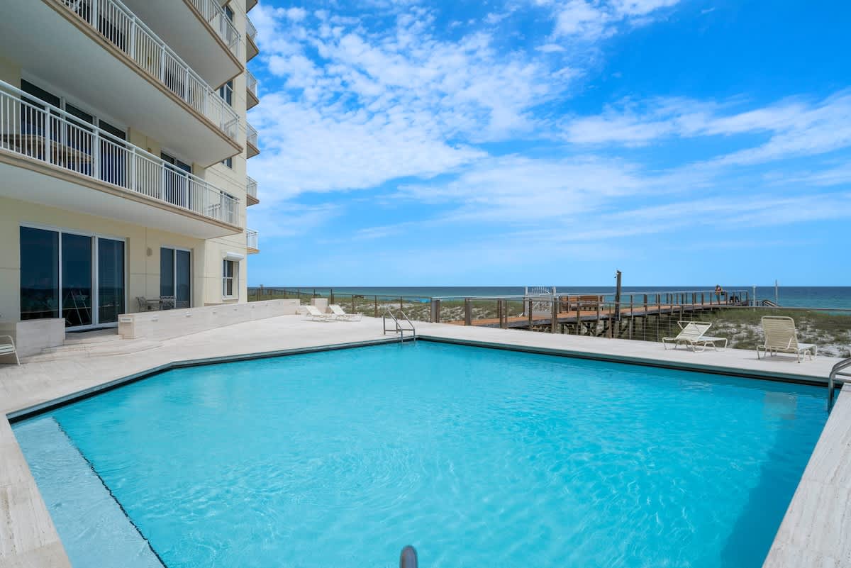 A large outdoor swimming pool with steps, surrounded by a light-colored deck, with the multi-story building exterior on one side and the ocean stretching to the horizon on the other.