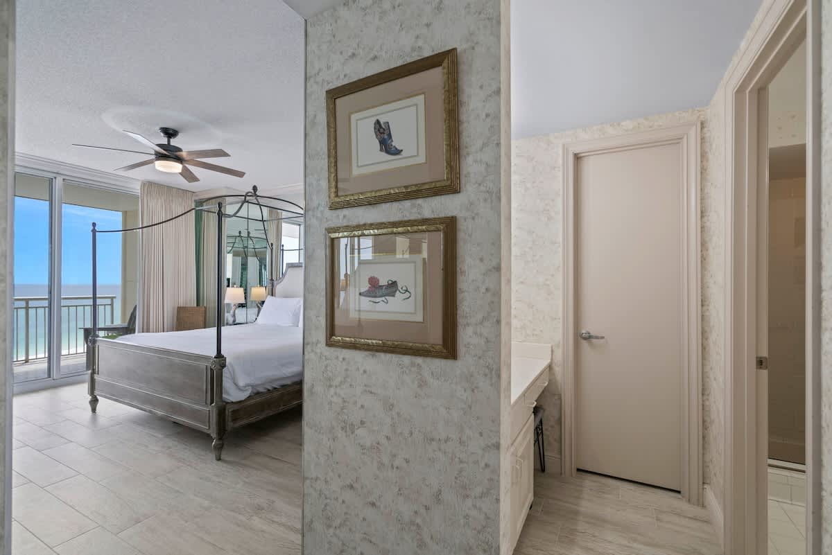A hallway with textured wallpaper leads into a bedroom with a four-poster bed and a balcony offering ocean views. Two framed art pieces hang on the wall.
