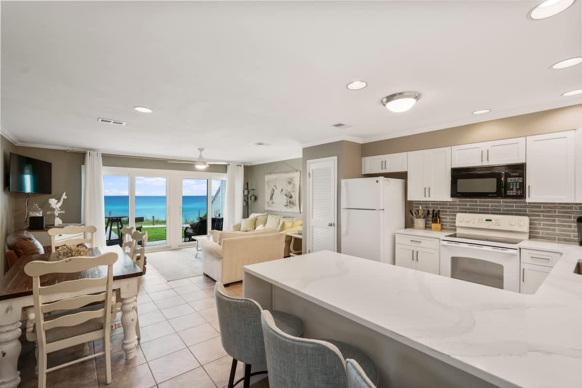 Open-concept living and dining area with a clear view of the ocean through large glass doors. The kitchen's breakfast bar with stools is visible on the right, connecting the spaces, all on light tile flooring.