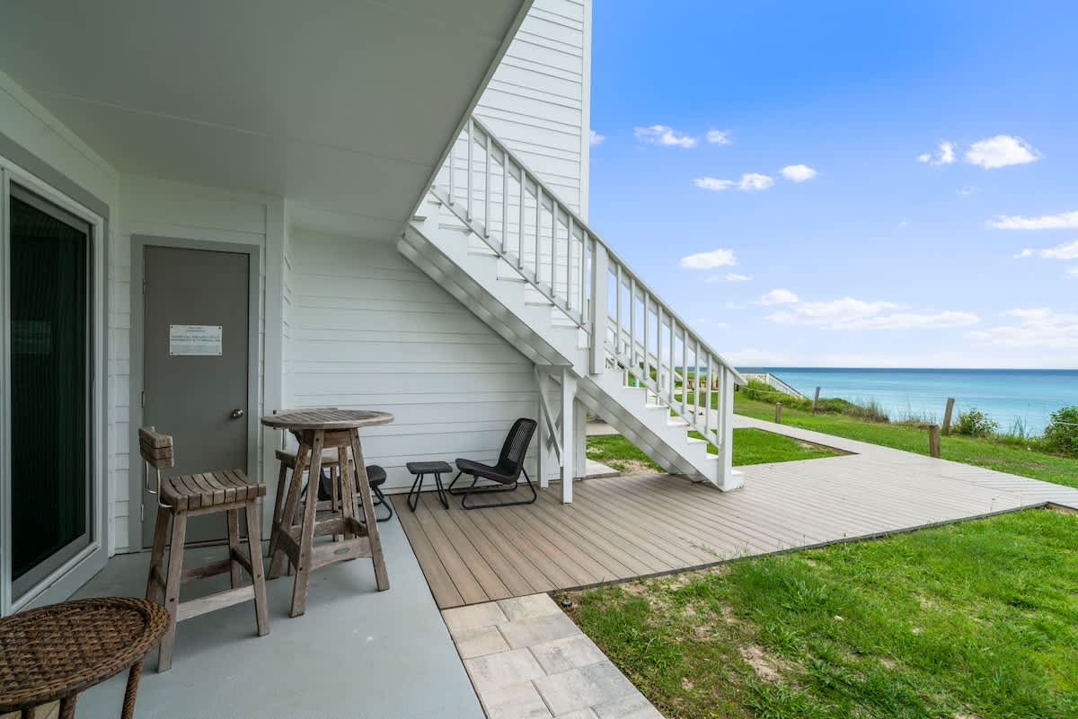 A ground-level patio area with a high-top table and stools, featuring a white staircase leading to an upper level, and expansive views of the ocean and coastline.