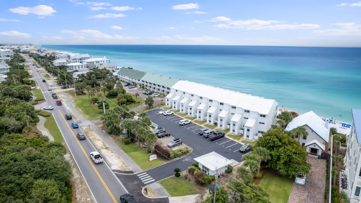 A wide aerial shot reveals a long stretch of coastline with a coastal road running parallel to the vibrant turquoise ocean, featuring a row of white townhouses and other buildings. The property's swimming pool and parking area are visible in the foreground.