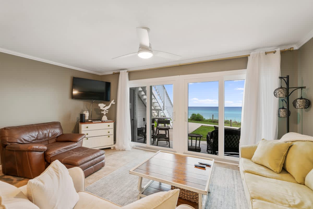 Spacious living room with a brown leather armchair and ottoman, light-colored sofas, and a wooden coffee table on a grey area rug. Floor-to-ceiling glass doors open to a patio with ocean views, and a ceiling fan hangs overhead.