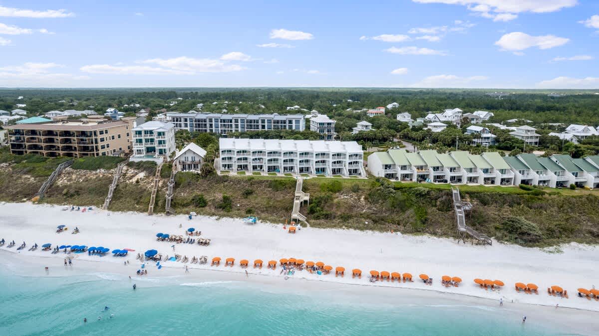 An aerial shot presents a vibrant white sand beach with clear turquoise waters, featuring rows of blue and orange beach setups. Behind the beach, a series of white and green-roofed townhouses and other multi-story buildings are nestled on a lush dune.