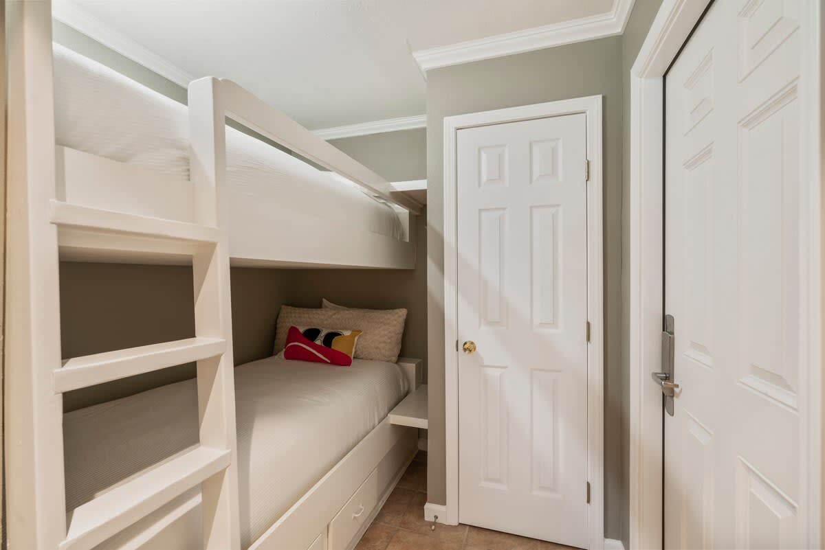 A cozy bunk nook features a built-in white wooden bunk bed with a ladder, striped bedding, and a colorful decorative pillow, adjacent to a white paneled door.