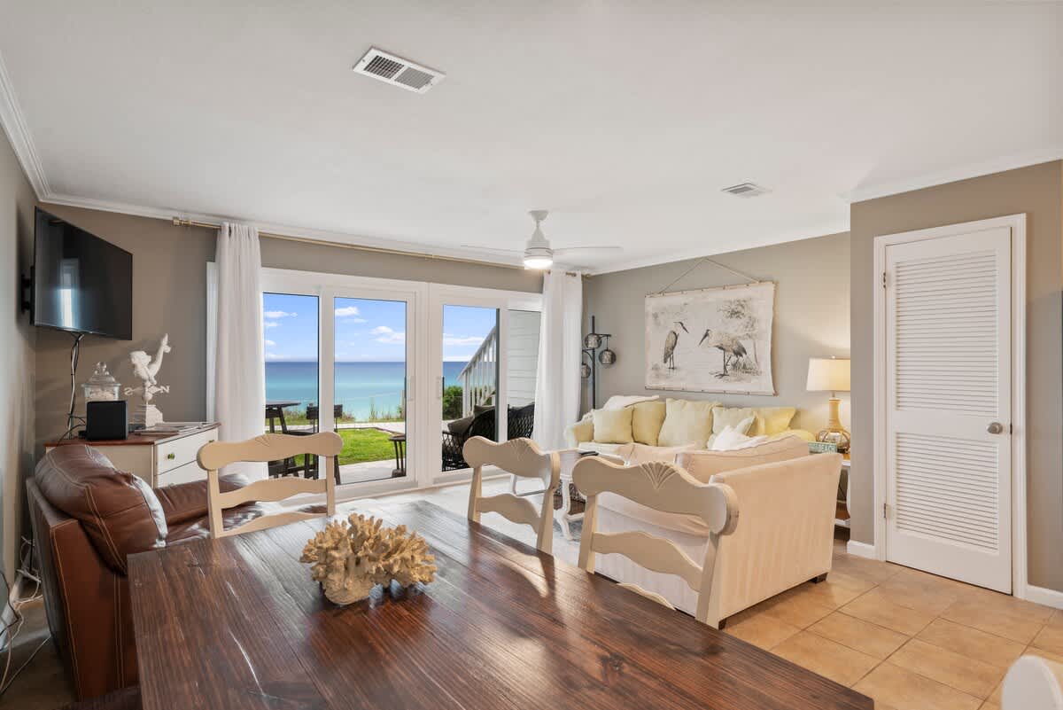 Open-concept living and dining area with a view of the ocean through large glass doors. The living space has light sofas, while the dining area features a dark wood table with light-colored chairs, all on tile flooring.