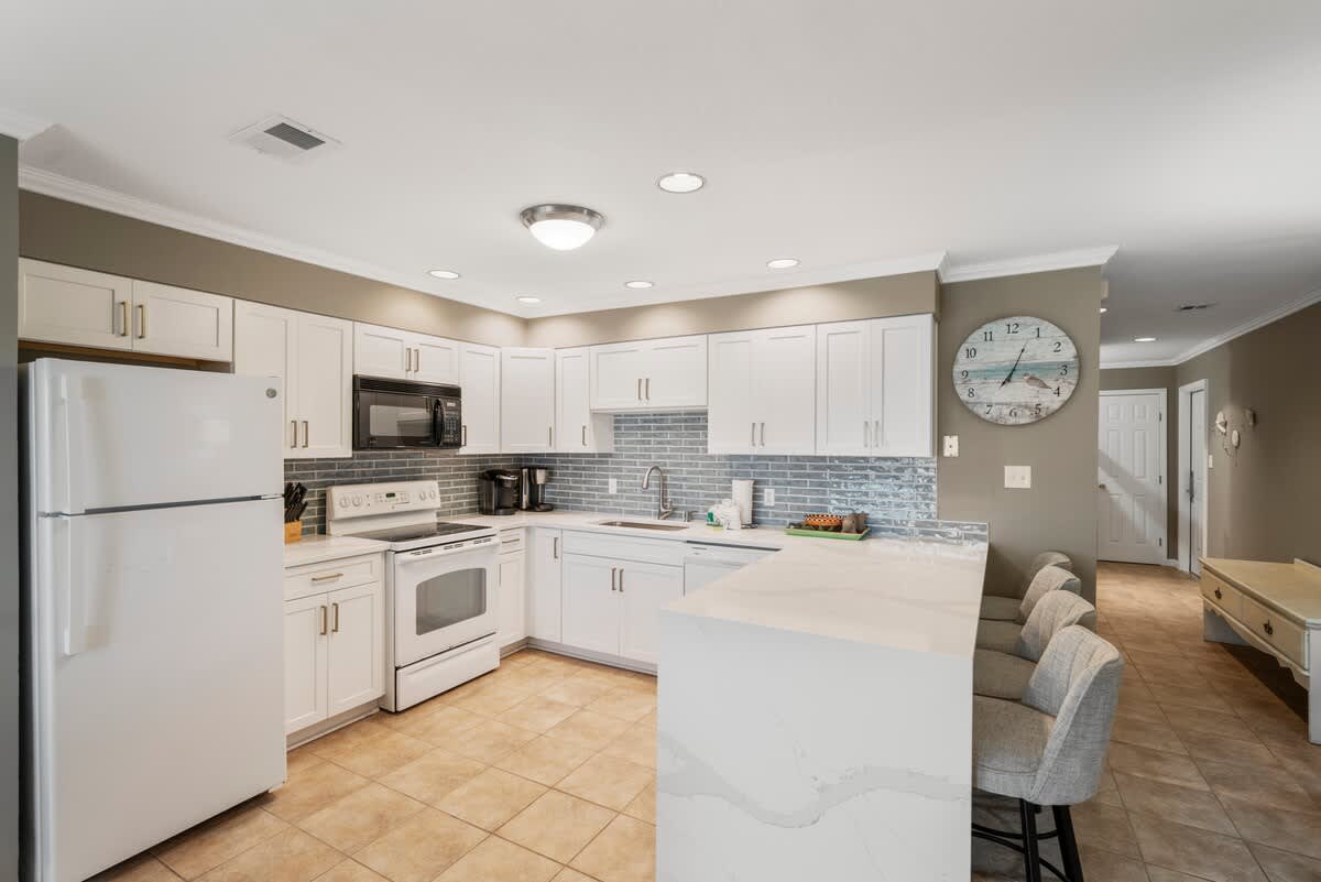 Spacious kitchen featuring white cabinets, light quartz countertops, and a blue subway tile backsplash. A white refrigerator and electric range are visible, along with a breakfast bar with three grey upholstered stools.