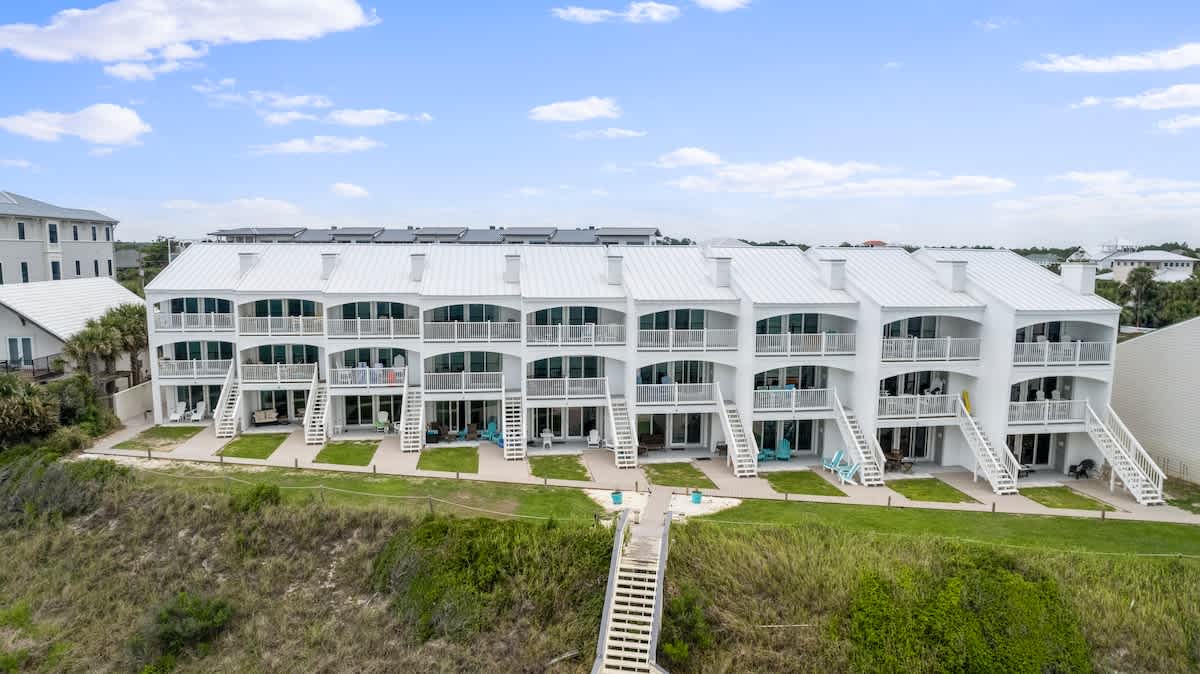 An aerial view focuses on the rear of a row of modern white townhouses, each featuring multiple balconies with white railings and ground-level patios, overlooking a grassy dune with wooden stairs leading to the beach.