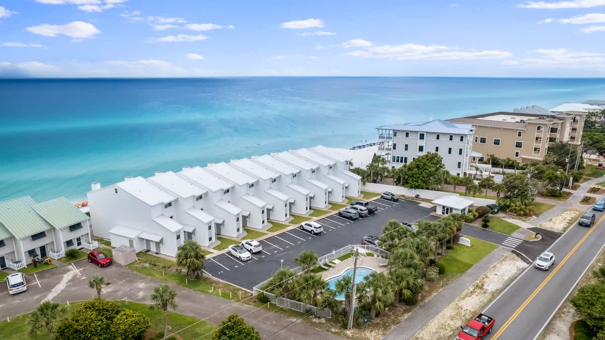 An aerial perspective captures a long row of white townhouses with a parking lot, positioned between a coastal road and the stunning turquoise ocean. A small, fenced swimming pool area is visible near the road, surrounded by palm trees.