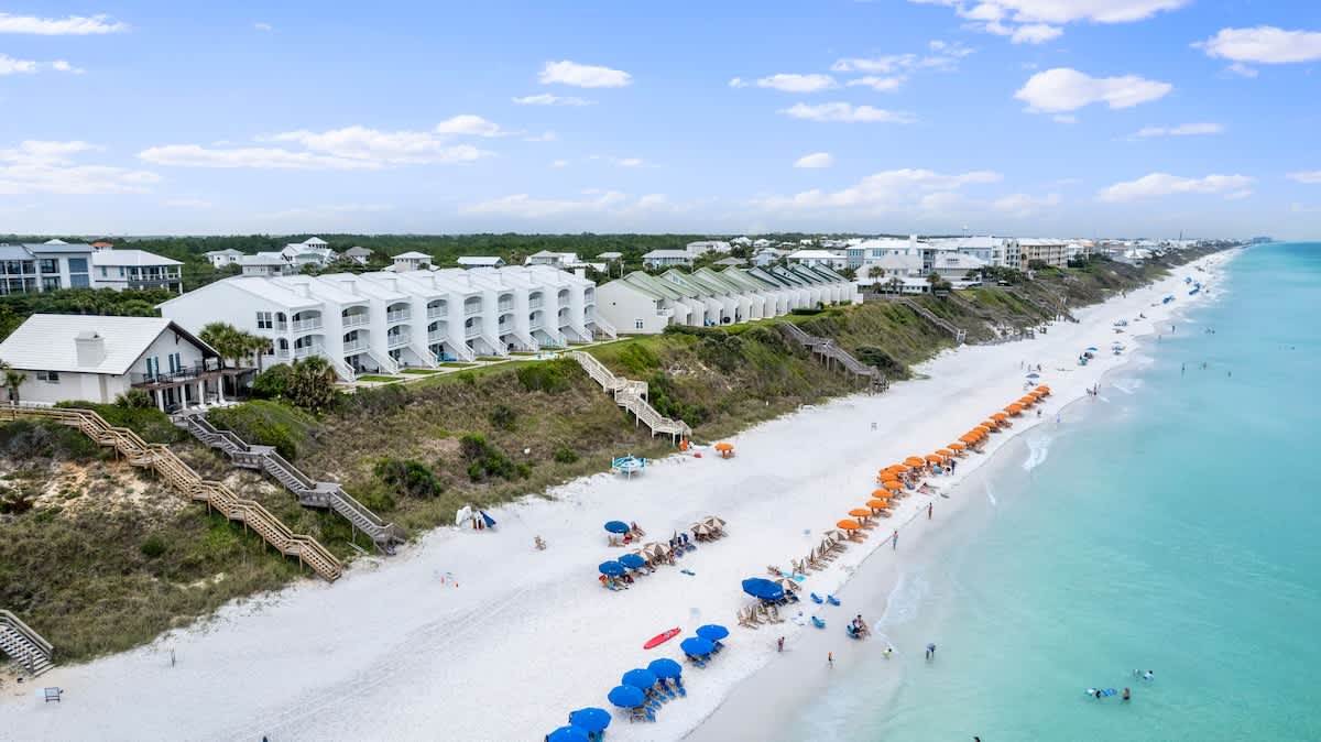 An aerial view captures a pristine white sand beach lined with numerous blue and orange beach umbrellas and chairs, with the turquoise ocean stretching into the horizon. A row of white and green-roofed townhouses sits atop a grassy dune overlooking the beach.