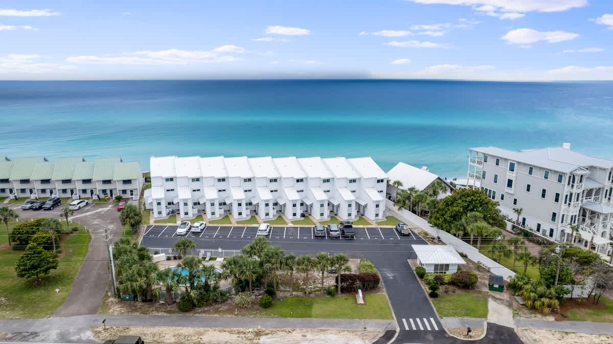 An aerial view showcases a row of white multi-story townhouses with parking in front, situated near a vibrant turquoise ocean under a partly cloudy sky. A small swimming pool area with palm trees is visible in the foreground.