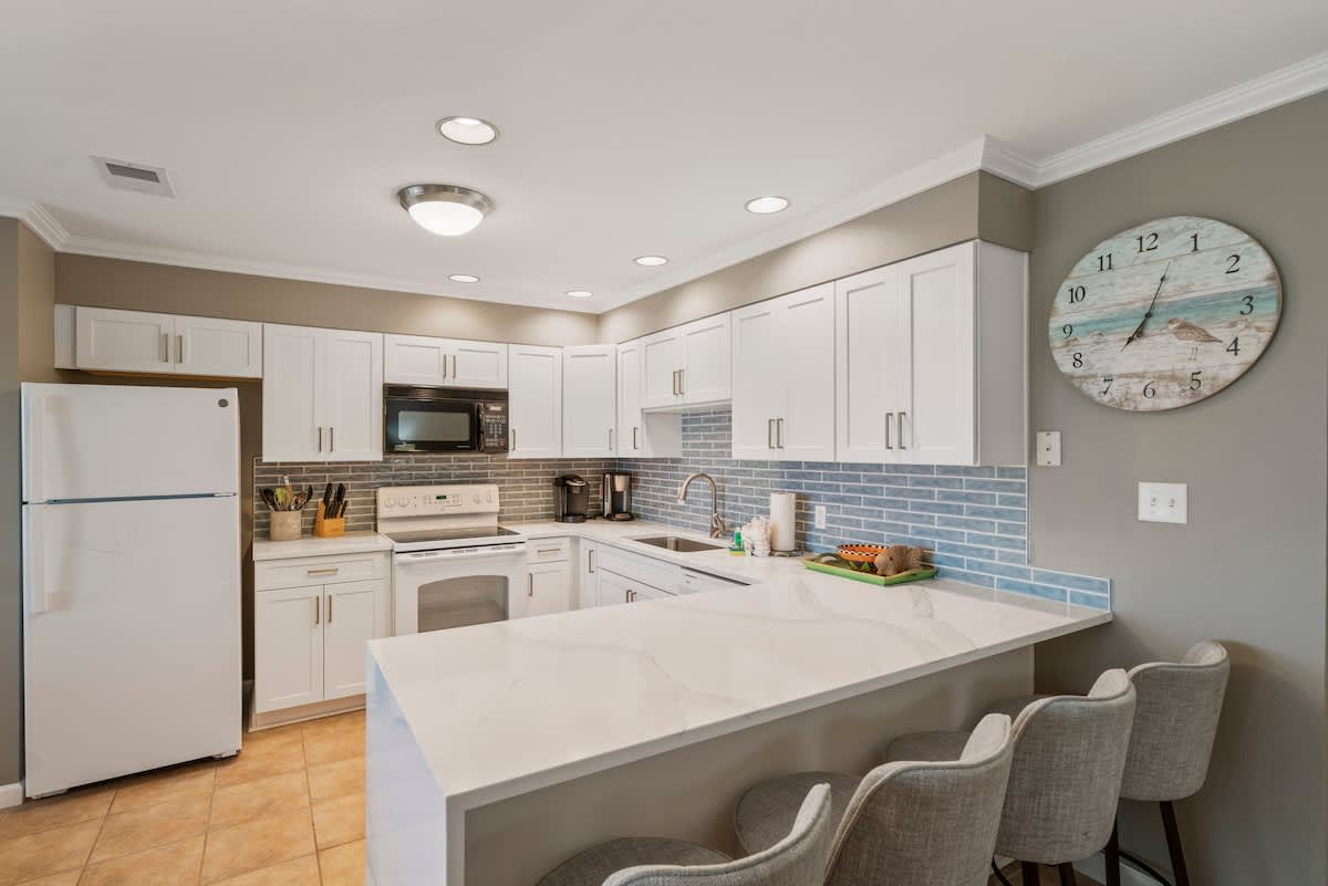 Well-lit kitchen with white shaker cabinets, light quartz countertops, and a blue subway tile backsplash. The kitchen includes a white refrigerator, electric range, and a breakfast bar with three grey stools, all on tile flooring.