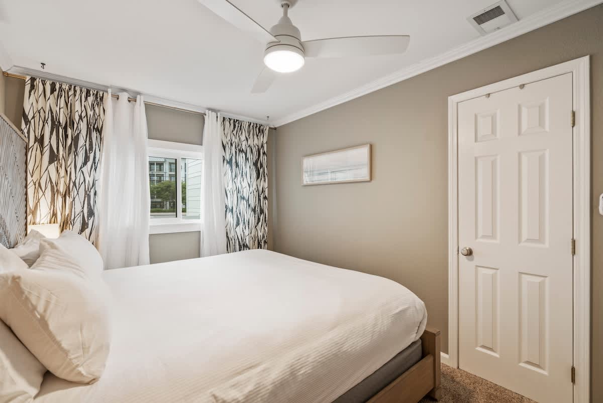 Corner view of the master bedroom, showing the king bed, a window with patterned curtains, and a white paneled door, all under a modern ceiling fan.