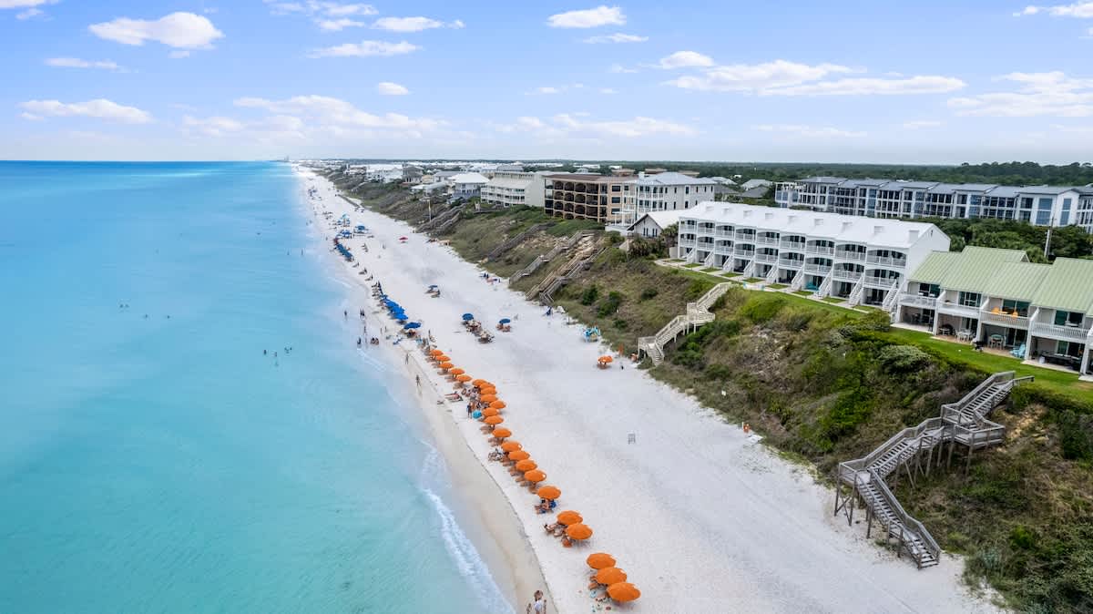 A sweeping aerial view showcases a vast expanse of white sand beach with turquoise ocean waters, dotted with colorful beach umbrellas and sunbathers. A long line of coastal properties, including white and green-roofed townhouses, are perched on a vegetated dune.