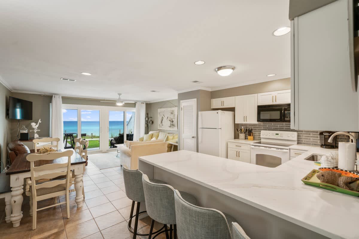 Panoramic view of the open-concept living, dining, and kitchen areas, all featuring light tile flooring. Large glass doors frame the ocean view, while the kitchen's breakfast bar and the dining table are visible in the foreground.