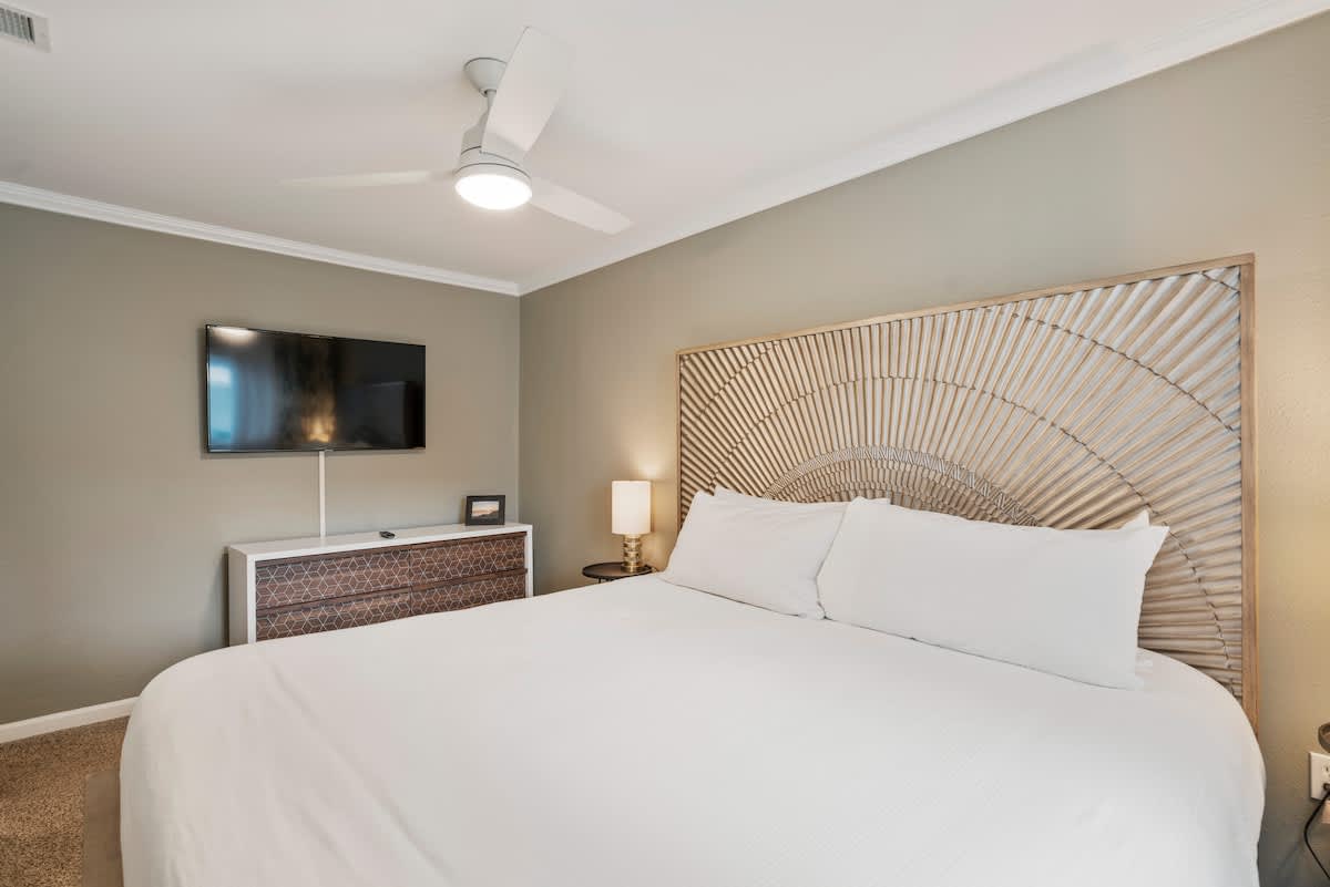A wide shot of the master bedroom showcasing the king bed with a distinctive headboard, a wall-mounted flat-screen TV above a dresser, and a modern ceiling fan.