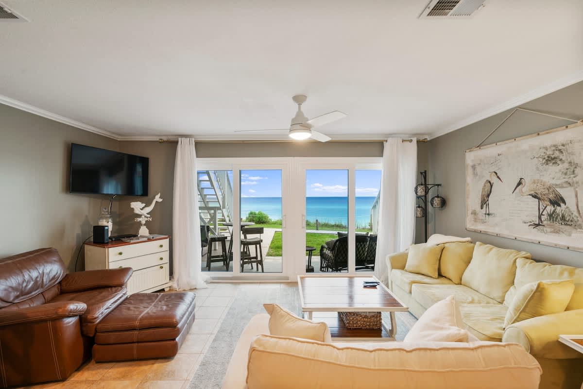 Coastal-themed living room featuring a brown leather armchair, light yellow sofas, and a wall-mounted TV. Large glass doors provide a stunning view of the ocean, with a patio and outdoor seating visible beyond.