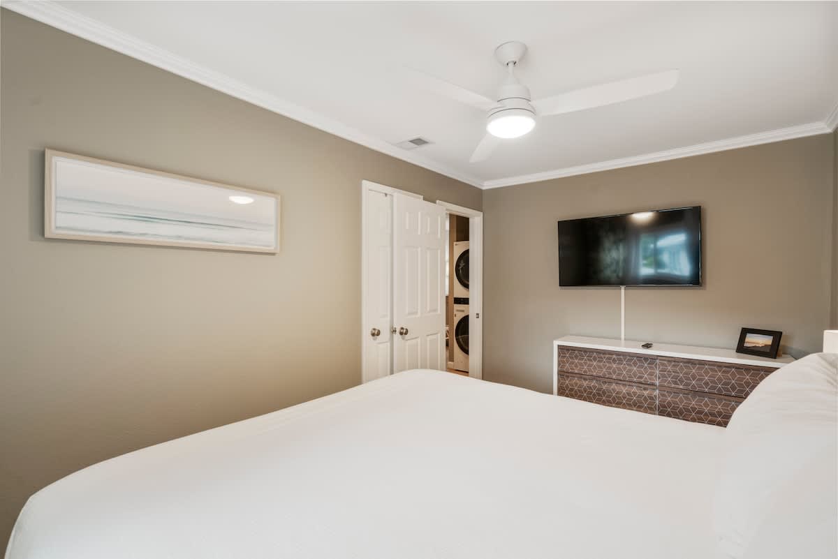View of the master bedroom from the foot of the bed, highlighting the wall-mounted TV, a dresser with geometric patterns, and a closet with bi-fold doors.