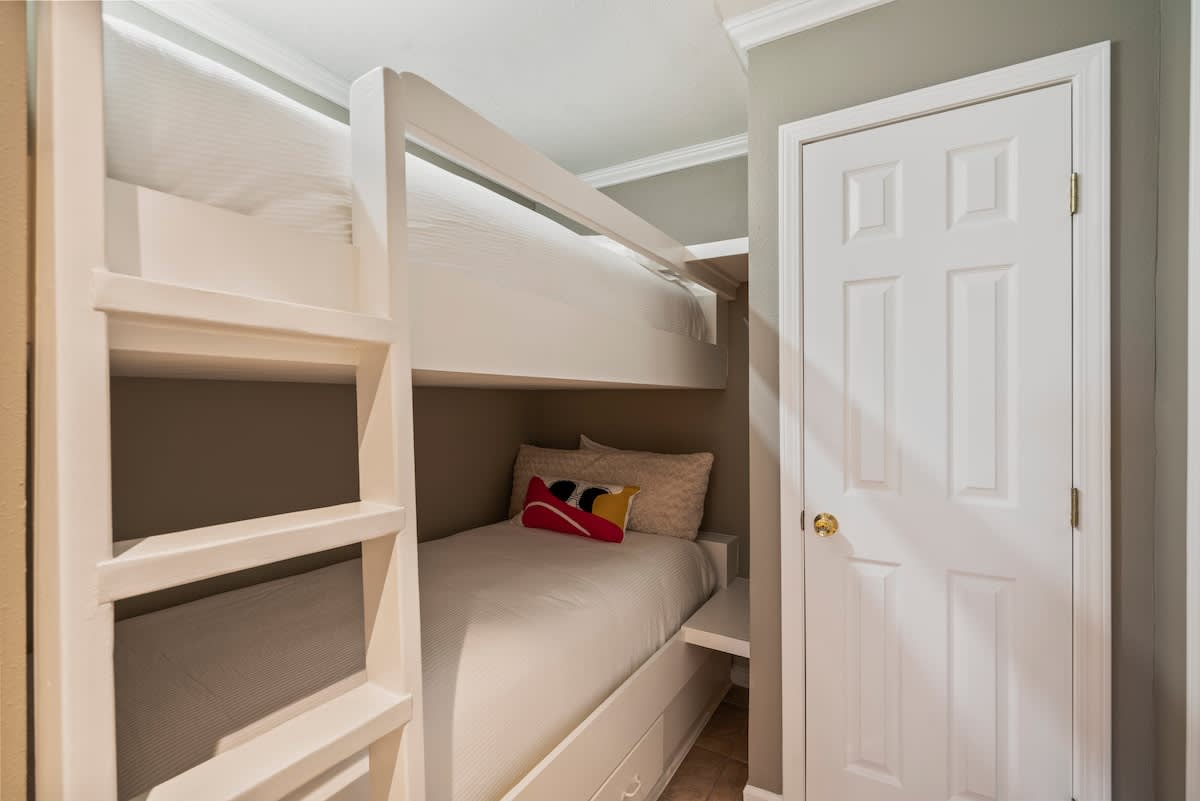 A close-up view of the built-in white wooden bunk bed, showing the lower bunk with a striped comforter and a vibrant throw pillow, next to a white paneled door.