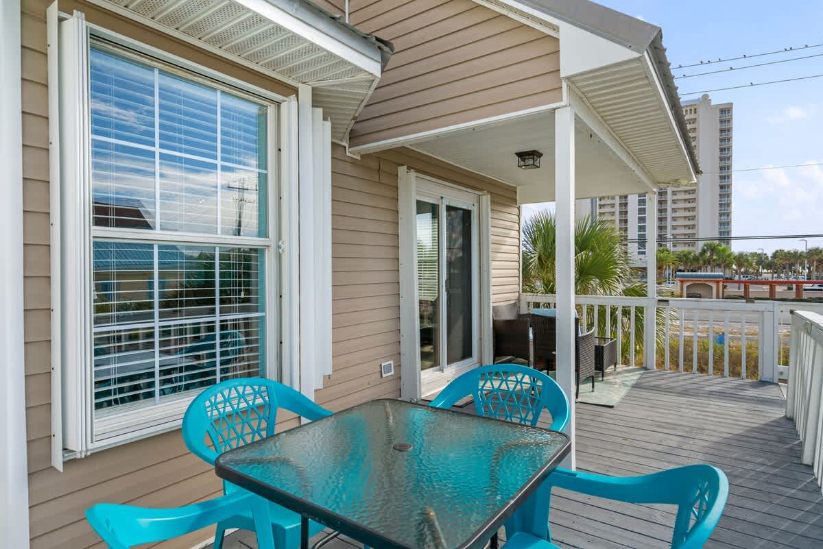 An outdoor deck features a glass-top table with four turquoise plastic chairs, set against a light brown siding house with a large window and sliding glass door, offering a glimpse of distant buildings.