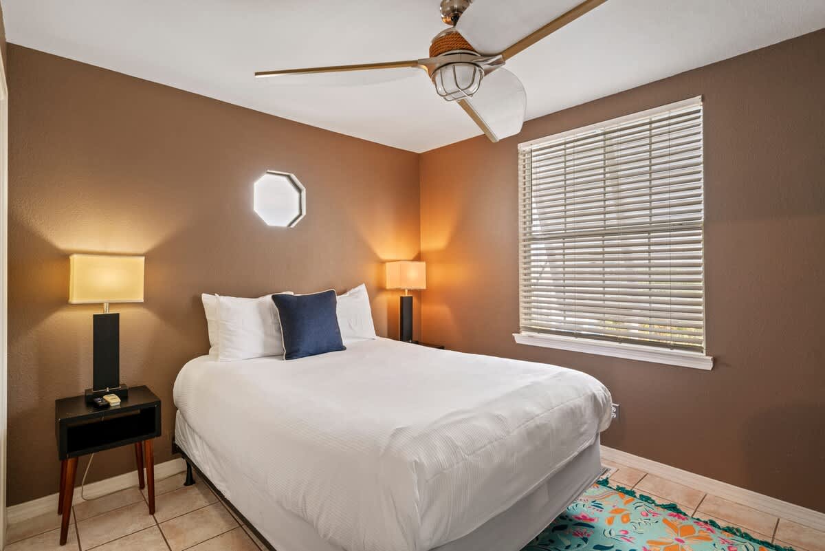 A cozy bedroom features a queen-sized bed with white linens and a blue accent pillow, flanked by two dark wood nightstands with lamps, under a ceiling fan, with a window covered by blinds and a colorful rug on tiled floor.