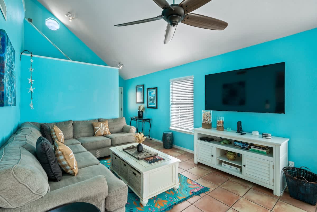 Cozy living room with bright turquoise walls, terra cotta tile floors, a large L-shaped sectional sofa, a white entertainment console with a flat-screen TV, and a window with blinds.