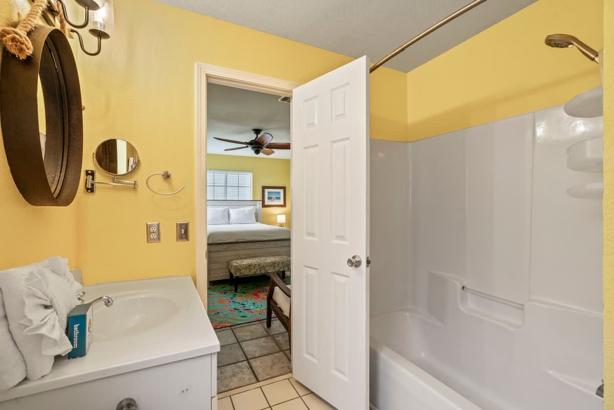 Sunny bathroom with yellow walls, a white vanity, and a shower/tub combo, with an open door revealing a glimpse into an adjacent bedroom with a bed and ceiling fan.