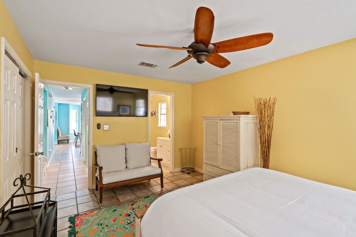 Spacious bedroom with yellow walls, a white bed, a wooden ceiling fan, and a wall-mounted TV, with a view into a hallway leading to a room with blue walls.