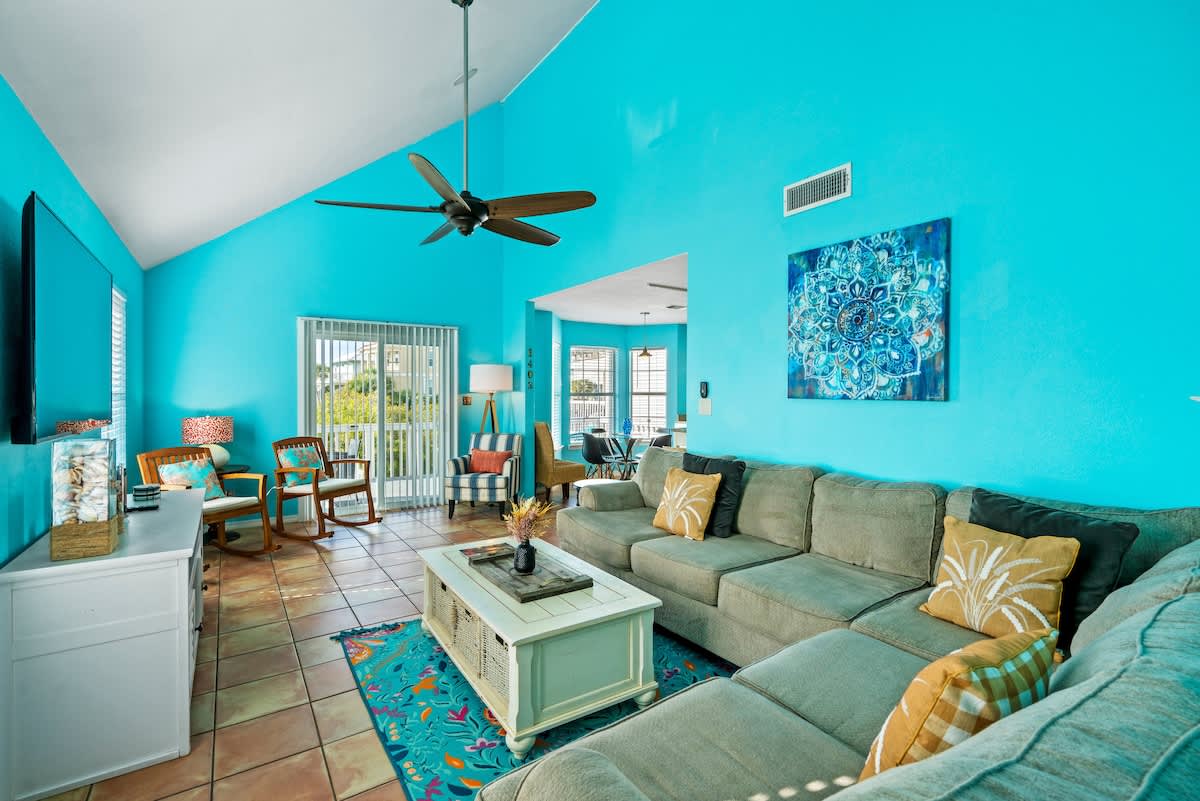 Expansive living room with vaulted ceilings and vibrant turquoise walls, featuring a large sectional sofa, a white coffee table, and a view towards the dining area and sliding glass door, all on terra cotta tile floors.