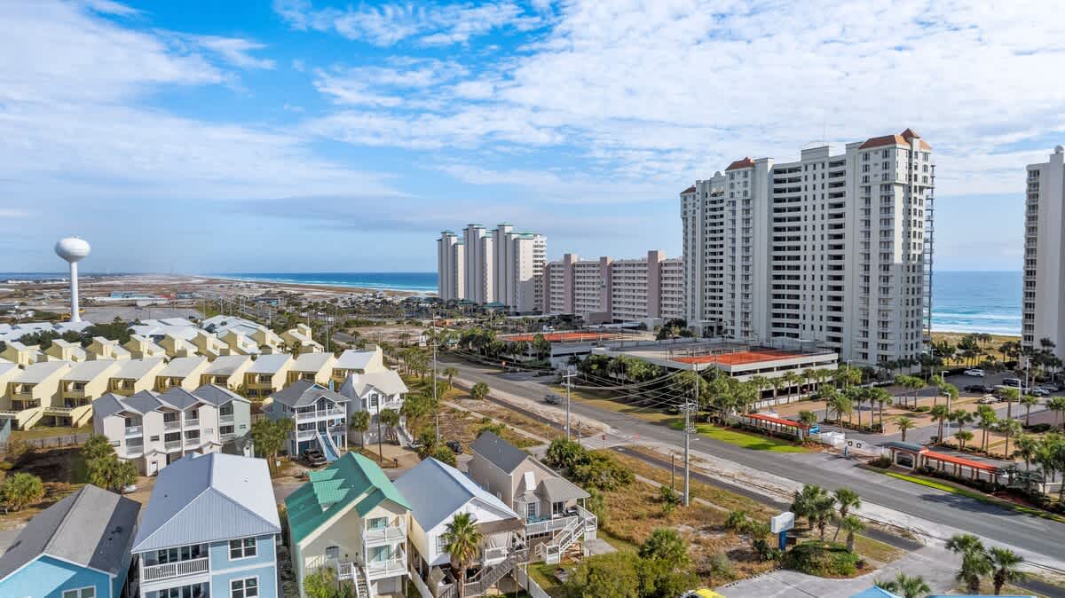 Aerial view of a coastal town with a mix of residential houses and tall condominium buildings, featuring a water tower, a road, and the ocean in the background under a partly cloudy sky.