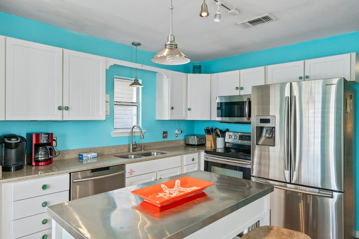 Detailed view of the kitchen showcasing white shaker cabinets, stainless steel appliances including a refrigerator, stove, and microwave, light granite countertops, and a double sink, all against bright turquoise walls.
