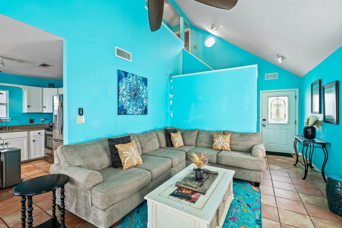 Open-concept living area featuring vibrant turquoise walls, terra cotta tile floors, a large L-shaped sectional sofa with decorative pillows, and a view into the adjacent kitchen with white cabinets.