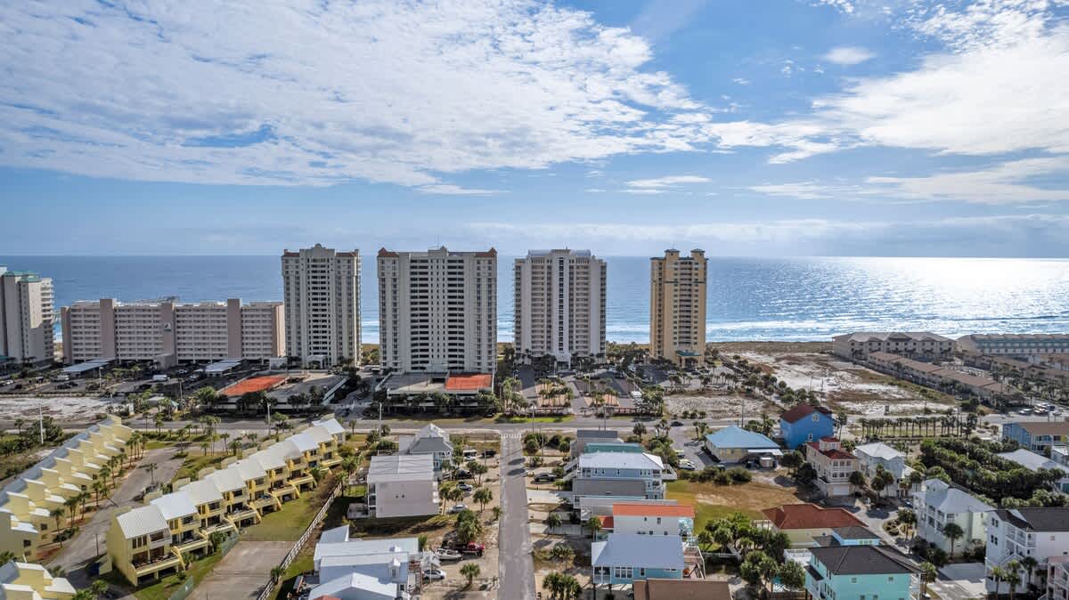 Expansive aerial view of a coastal landscape featuring multiple high-rise buildings along the beachfront, with residential homes and a road in the foreground, all under a bright blue sky with scattered clouds.