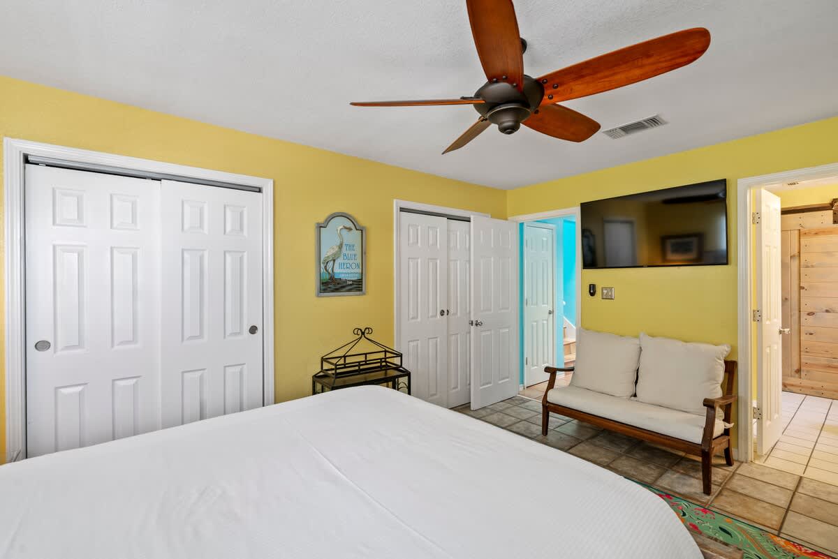 Cheerful bedroom with yellow walls, a white bed, a wooden ceiling fan, and a wall-mounted TV, featuring two white bi-fold closet doors and an open doorway to an adjacent room.