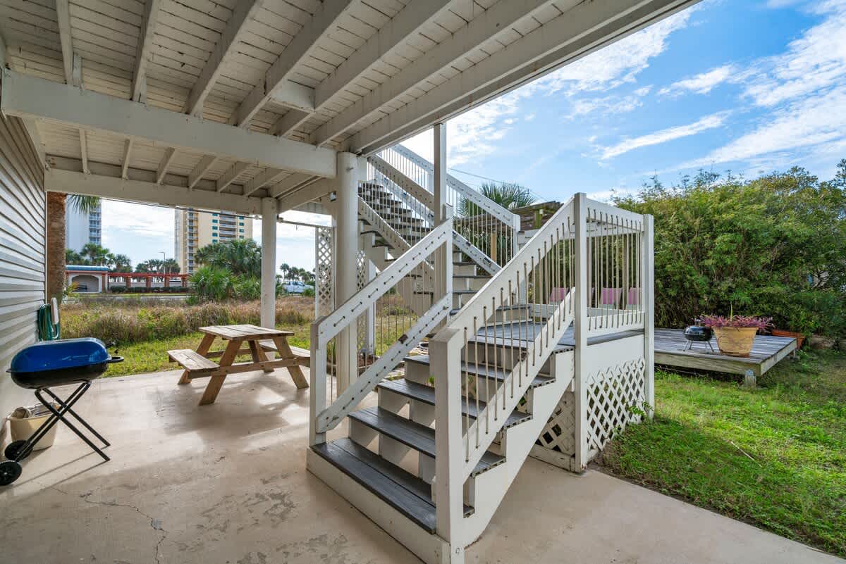 Shaded outdoor area beneath a raised house, featuring a concrete patio with a wooden picnic table and a BBQ grill, with white wooden stairs leading up to the main level.