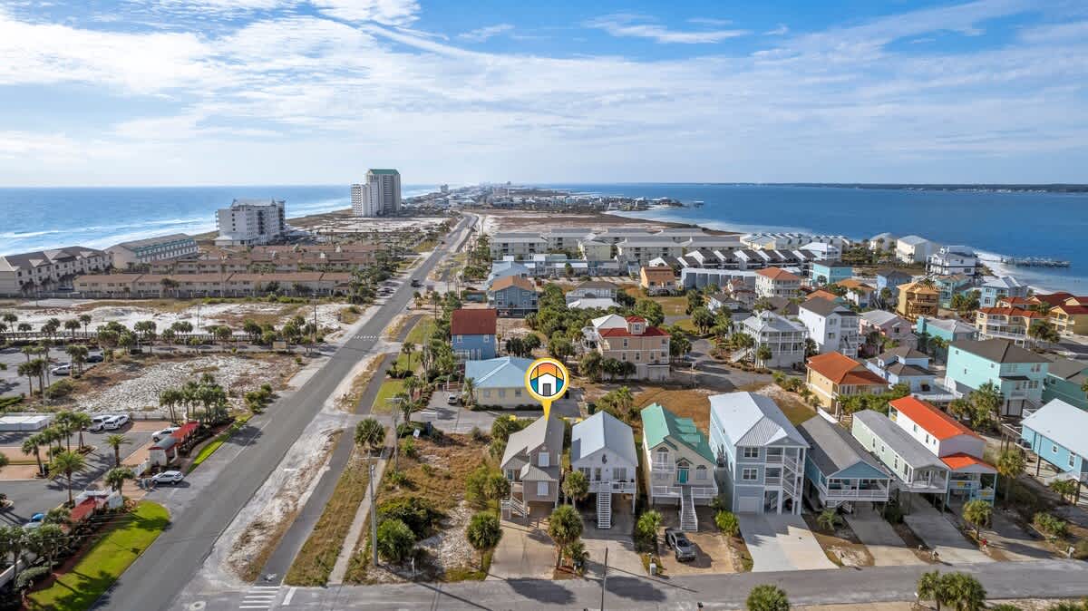 Wide aerial shot of a vibrant coastal town, featuring a mix of colorful residential houses and distant high-rise buildings, with the ocean and a bay visible, and a marker indicating the property.