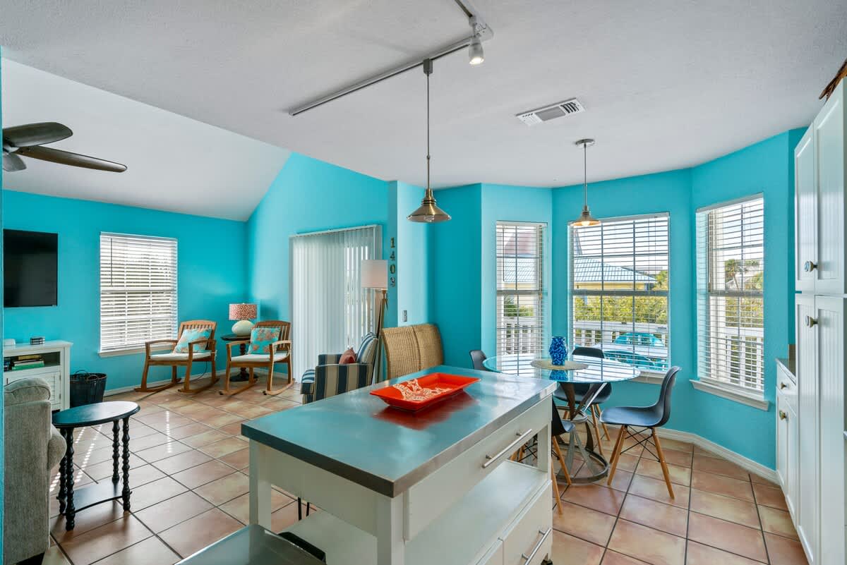 Open-plan living and dining area with bright turquoise walls and terra cotta tile floors, featuring a central kitchen island, a round glass dining table, and a view of the living room with a large TV and seating.