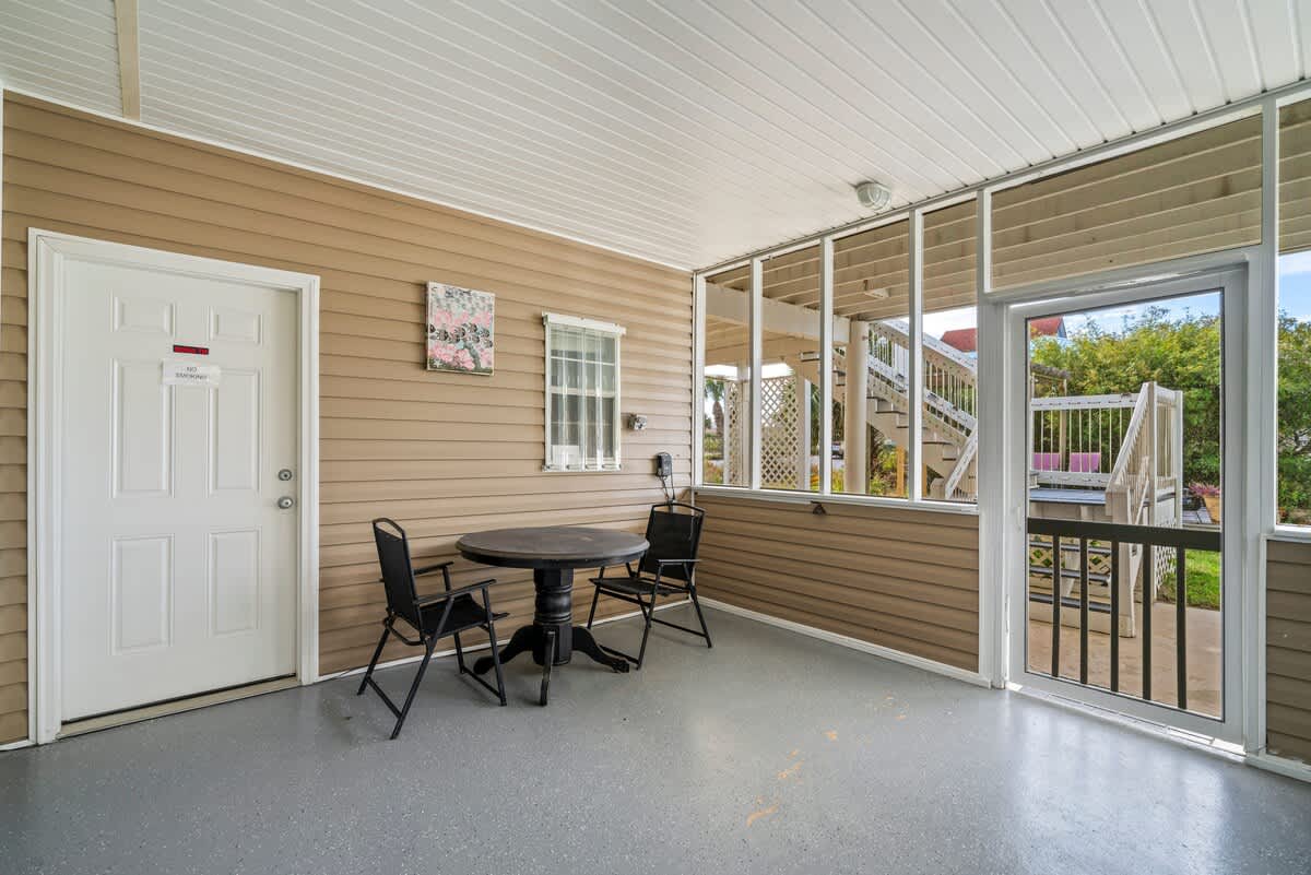 A covered and screened-in porch features a dark round table with two black chairs, set against light brown siding, with a white door on the left and a screened door leading to outdoor stairs on the right.